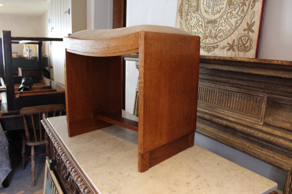 An oak stool featuring solid slab side supports, a central connecting stretcher, and a concave upholstered fabric seat cushion. View VI