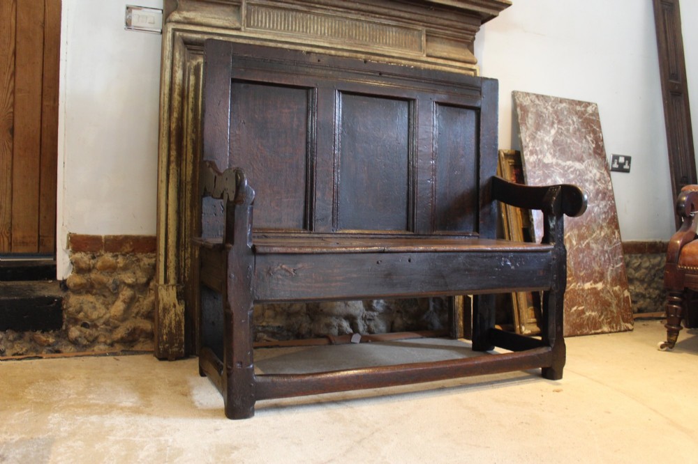 A dark oak 17th-century style settle bench with a three-panel backrest, shaped armrests, and wooden stretchers. View V