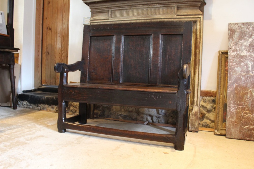 A dark oak 17th-century style settle bench with a three-panel backrest, shaped armrests, and wooden stretchers. View X