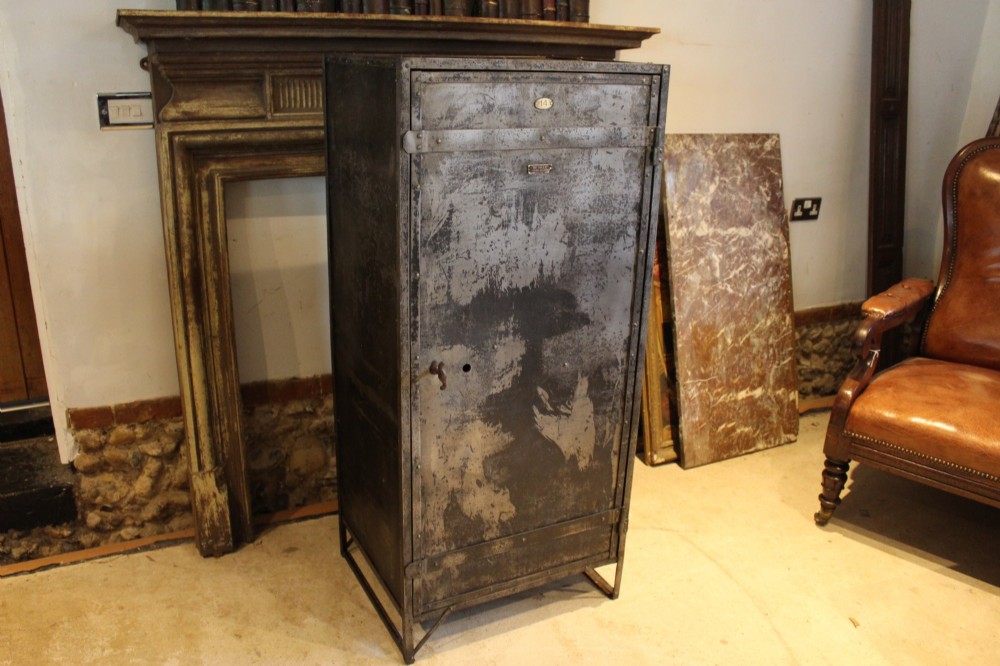 A tall, narrow Victorian industrial metal cupboard featuring a single door with a dark grey patinated surface, riveted frame, brass number plaque, and latch handle.