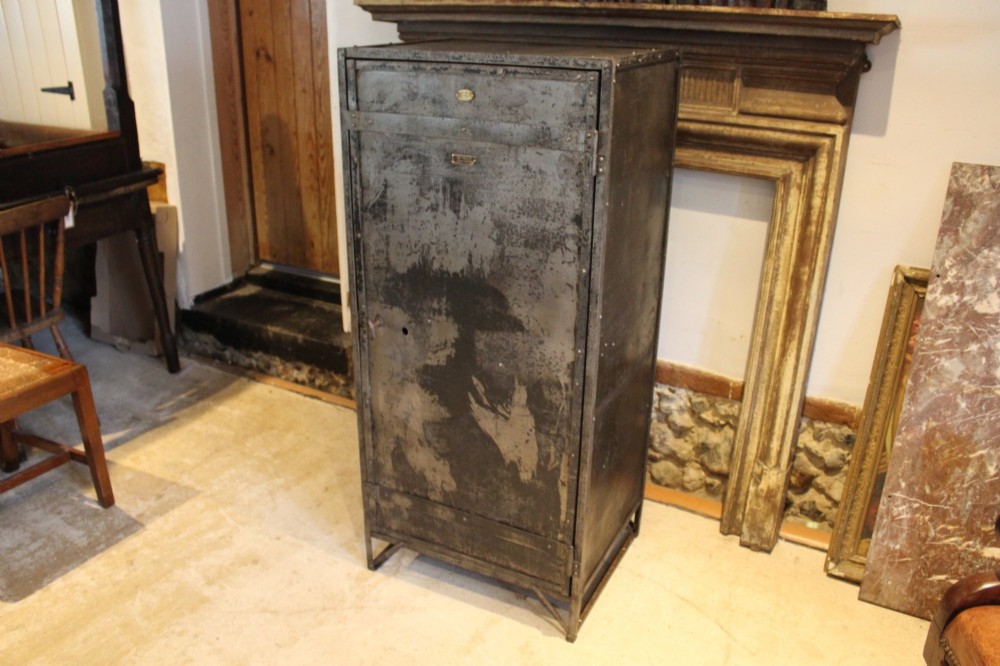A tall, narrow Victorian industrial metal cupboard featuring a single door with a dark grey patinated surface, riveted frame, brass number plaque, and latch handle. View X