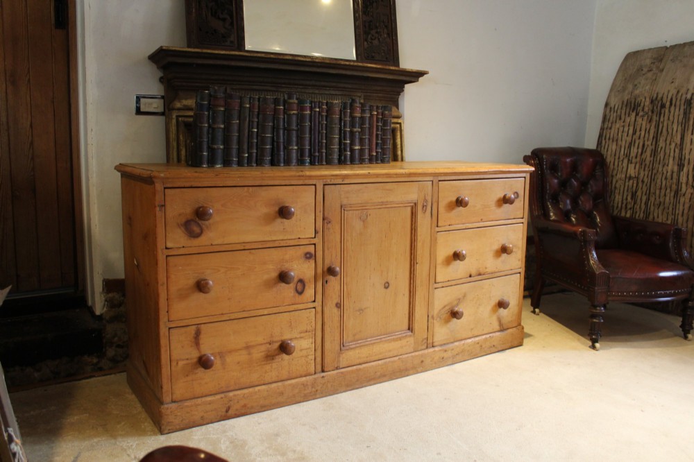 An early Victorian honey pine dresser base featuring a central panelled cupboard door flanked by three drawers on each side with turned wooden knobs.