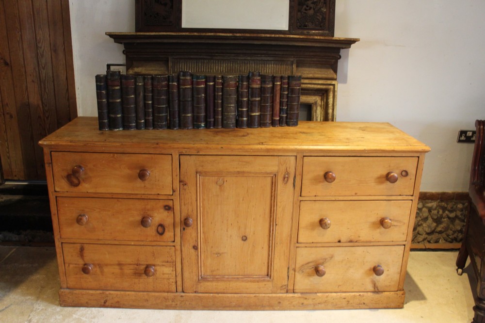 An early Victorian honey pine dresser base featuring a central panelled cupboard door flanked by three drawers on each side with turned wooden knobs. View II