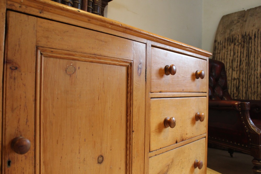 An early Victorian honey pine dresser base featuring a central panelled cupboard door flanked by three drawers on each side with turned wooden knobs. View III