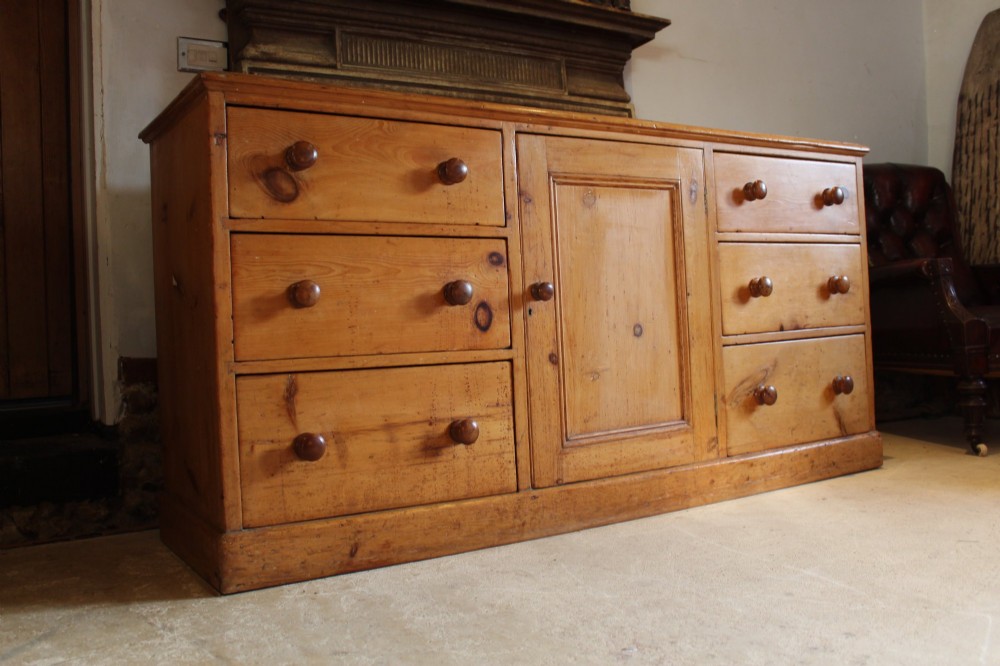 An early Victorian honey pine dresser base featuring a central panelled cupboard door flanked by three drawers on each side with turned wooden knobs. View VII