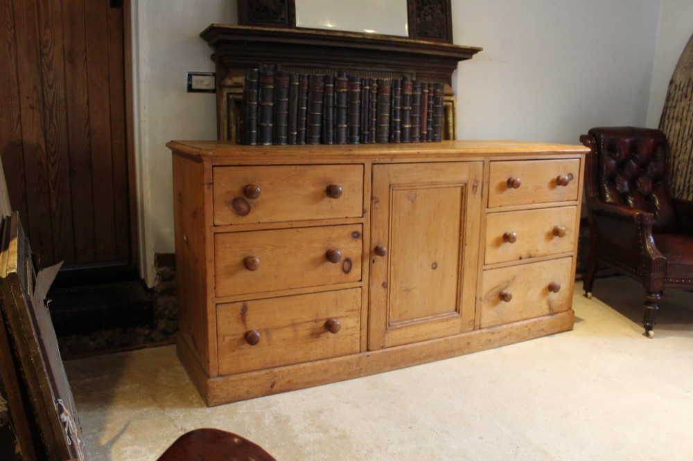 An early Victorian honey pine dresser base featuring a central panelled cupboard door flanked by three drawers on each side with turned wooden knobs. View VIII