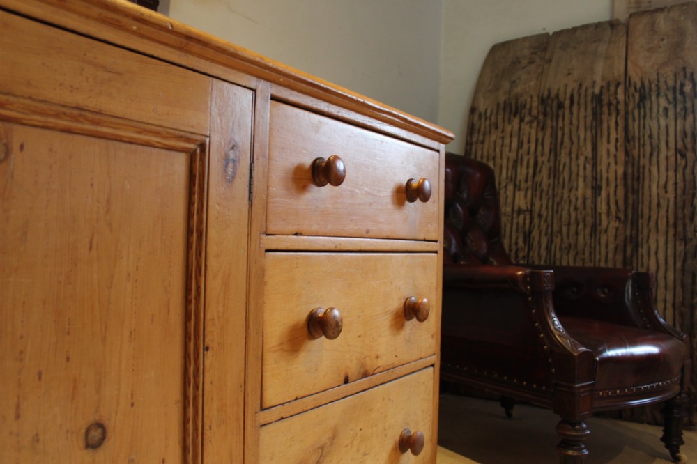 An early Victorian honey pine dresser base featuring a central panelled cupboard door flanked by three drawers on each side with turned wooden knobs. View IX