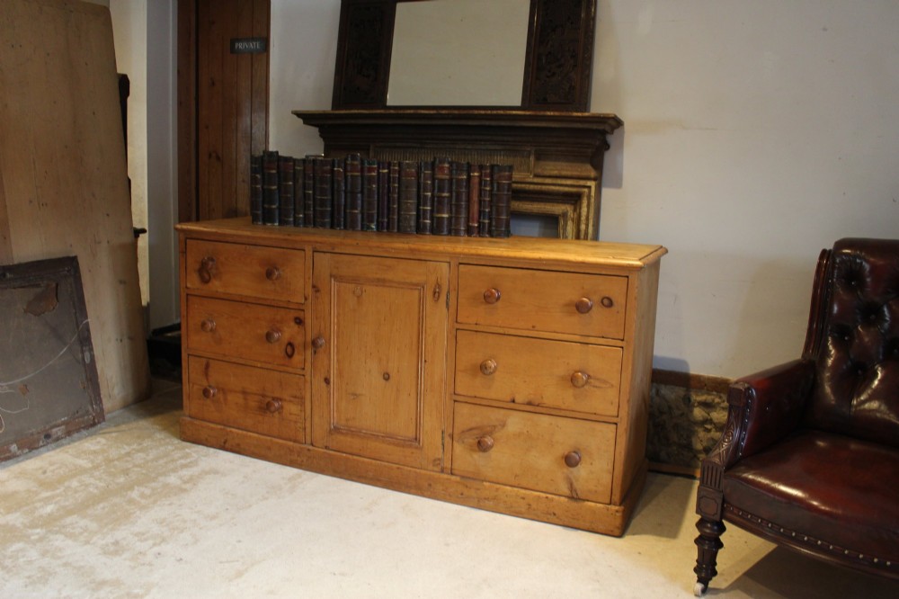 An early Victorian honey pine dresser base featuring a central panelled cupboard door flanked by three drawers on each side with turned wooden knobs. View XI