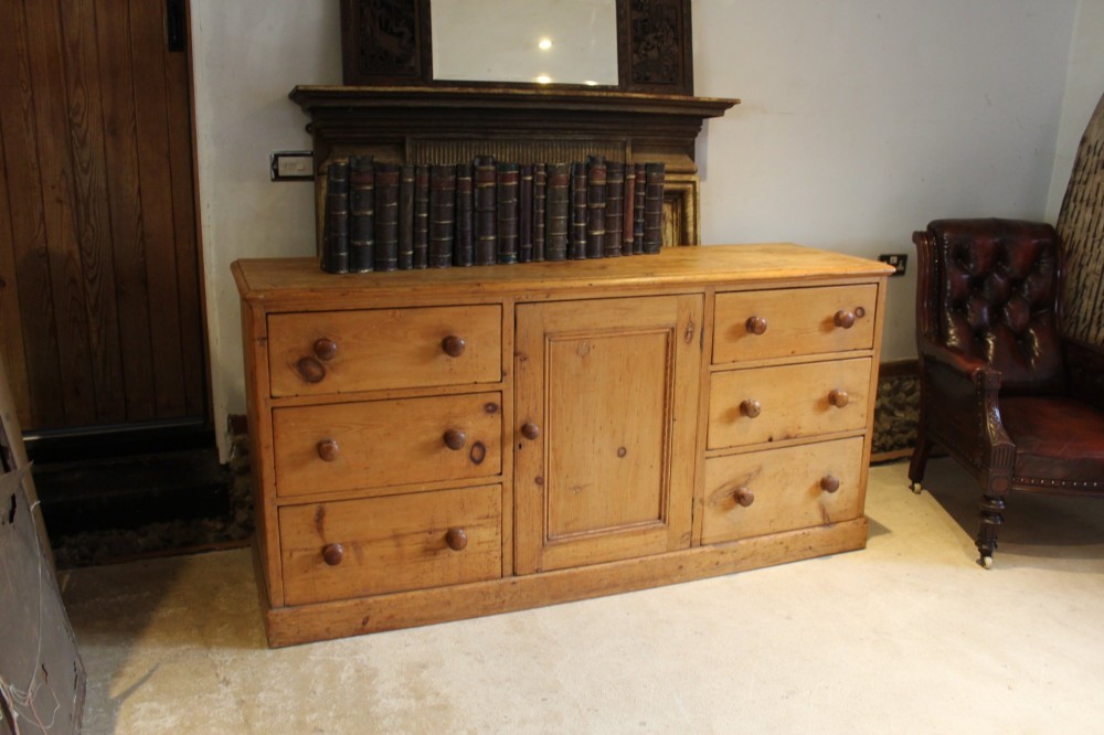 An early Victorian honey pine dresser base featuring a central panelled cupboard door flanked by three drawers on each side with turned wooden knobs. View XIII