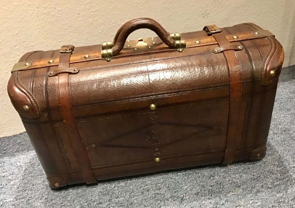 An antique dark brown leather valise suitcase featuring brass hardware, stitched straps, and embossed geometric detailing.