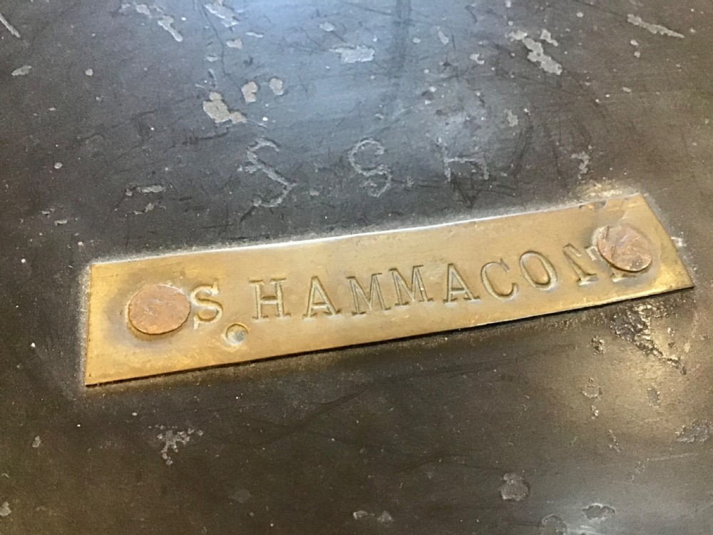A circular dark metal naval cap box with a hinged lid, front latch, and a riveted nameplate on top reading S. Hammacott. View III
