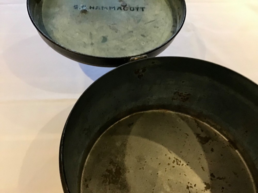 A circular dark metal naval cap box with a hinged lid, front latch, and a riveted nameplate on top reading S. Hammacott. View IV