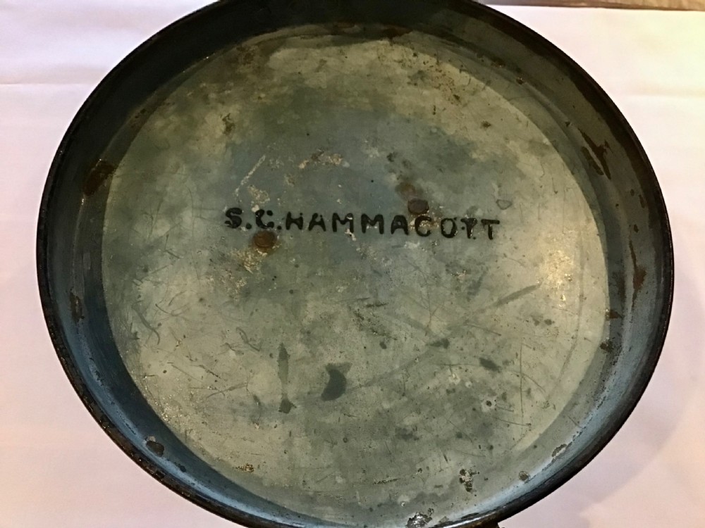 A circular dark metal naval cap box with a hinged lid, front latch, and a riveted nameplate on top reading S. Hammacott. View VI