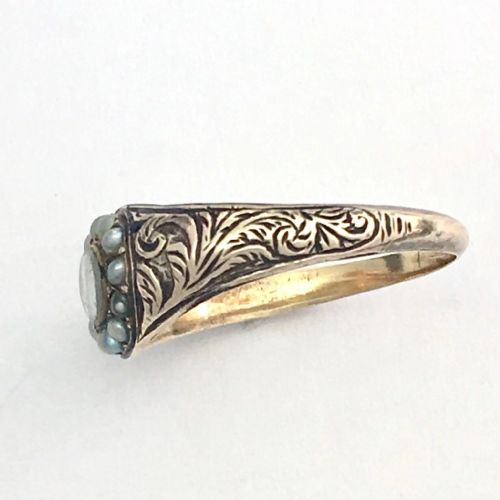 fig 1. A Georgian 9ct gold mourning ring featuring a central oval glass compartment containing hair surrounded by a halo of seed pearls and scroll-engraved shoulders.