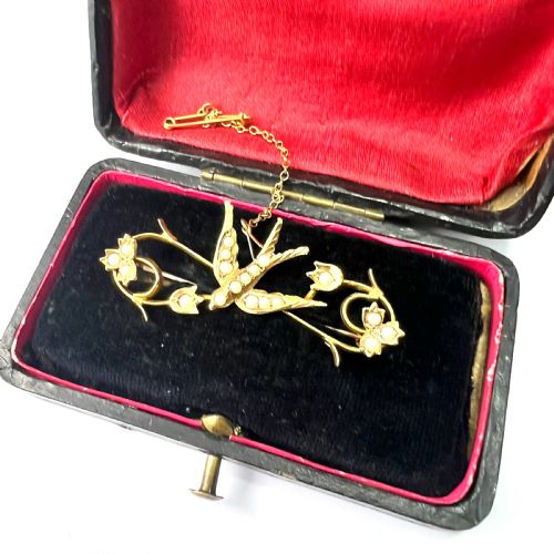 fig 1. An Edwardian 15ct yellow gold brooch featuring a central swallow motif set with seed pearls alongside floral scrollwork and a safety chain attachment.