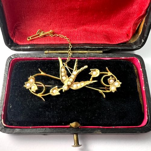 fig 2. An Edwardian 15ct yellow gold brooch featuring a central swallow motif set with seed pearls alongside floral scrollwork and a safety chain attachment.