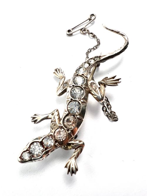 fig 1. An Art Deco silver brooch shaped like a lizard, encrusted with round clear paste stones along its spine and head, featuring an attached safety chain.