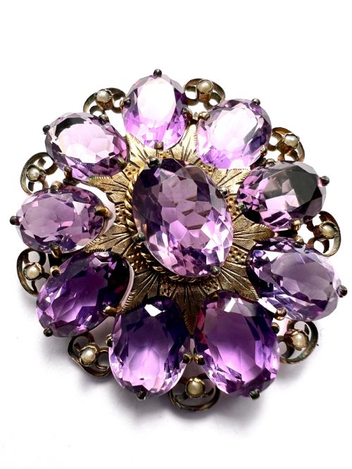fig 1. A circular Victorian gold brooch featuring a cluster of oval faceted amethysts around a central oval amethyst, bordered by textured gold leaf details and scrollwork set with seed pearls.