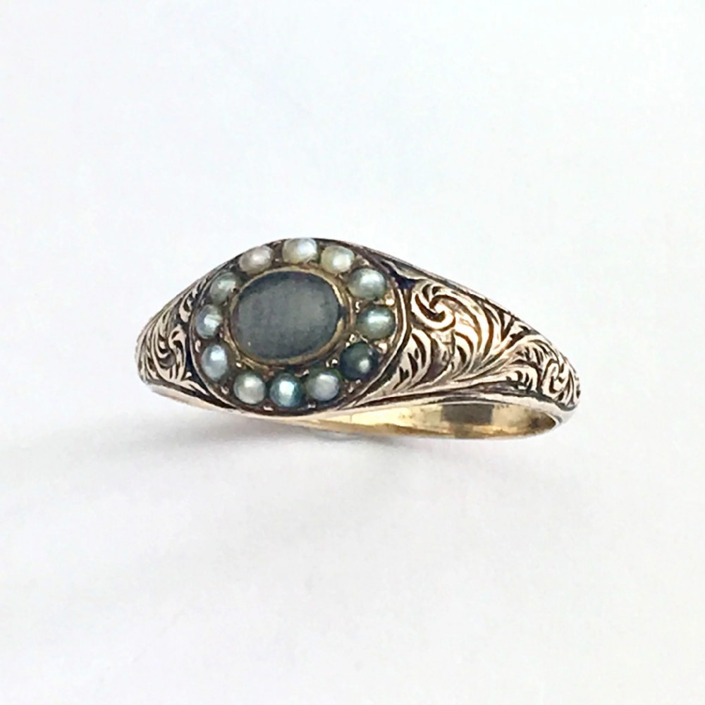 A Georgian 9ct gold mourning ring featuring a central oval glass compartment containing hair surrounded by a halo of seed pearls and scroll-engraved shoulders.