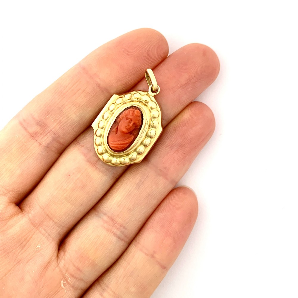 An oval Victorian 18ct gold pendant featuring an orange-red carved coral cameo classical bust framed by a beaded gold border. View V
