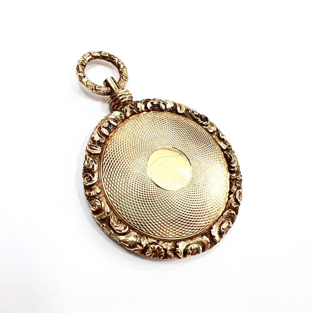 A Victorian 15ct yellow gold oval locket pendant featuring an engine-turned centre, a blank central cartouche, and a chased floral border.