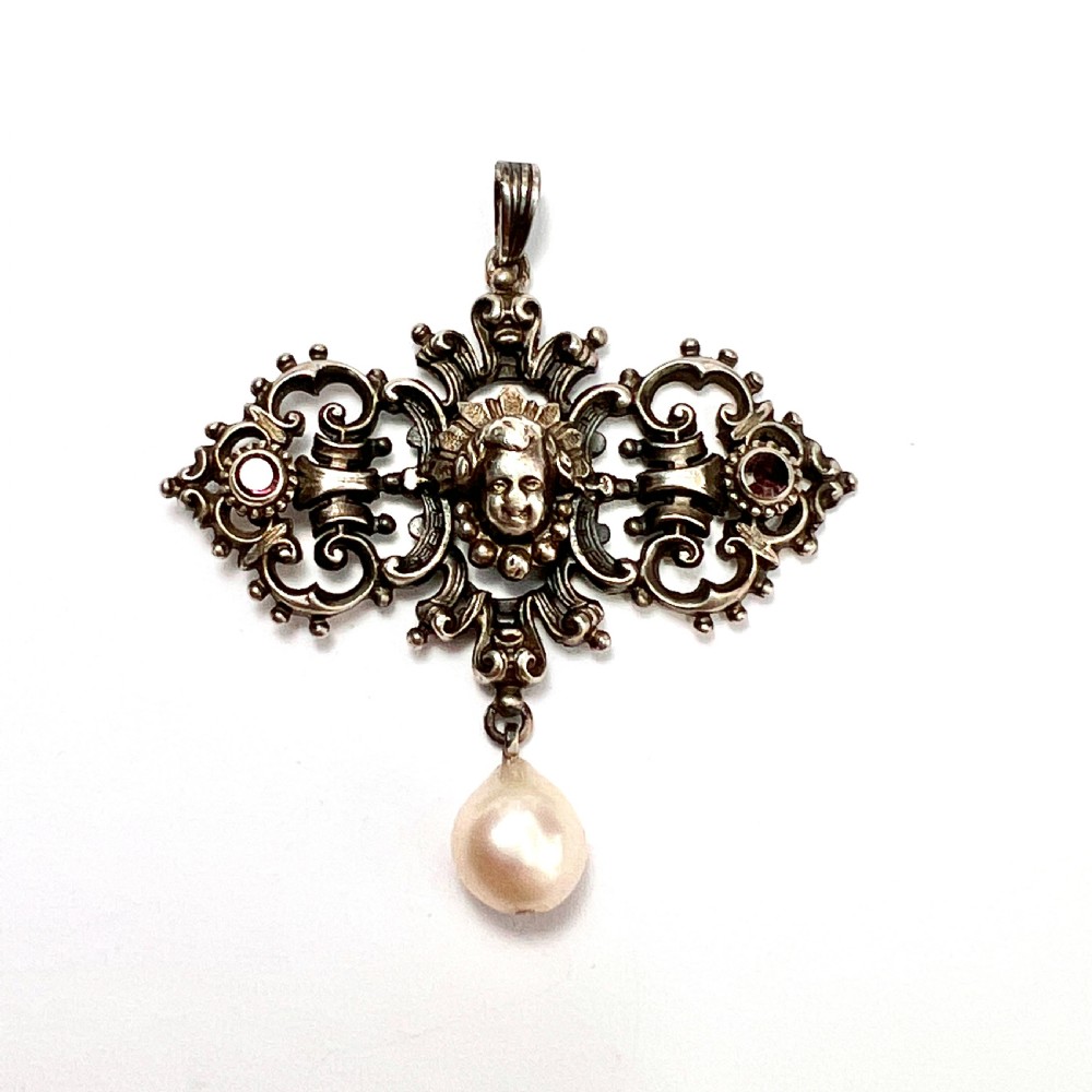 An antique silver pendant featuring scrollwork ornamentation, a central cherub mask, ruby-toned side accents, and a suspended baroque pearl drop.