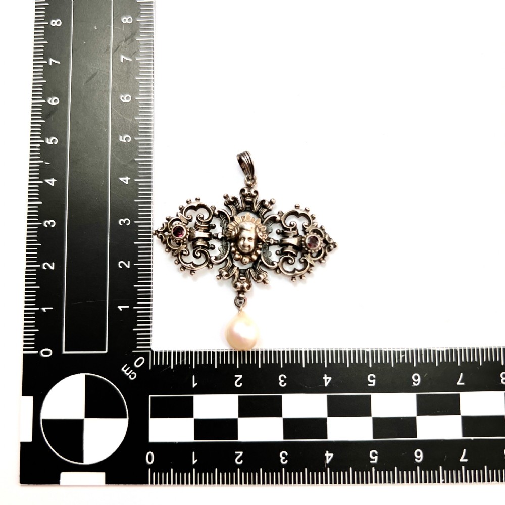 An antique silver pendant featuring scrollwork ornamentation, a central cherub mask, ruby-toned side accents, and a suspended baroque pearl drop. View II