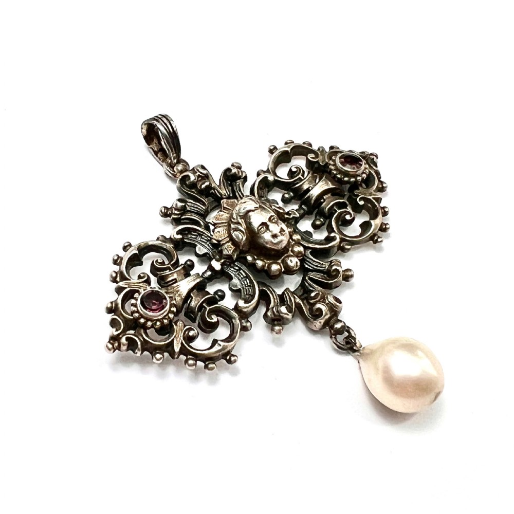 An antique silver pendant featuring scrollwork ornamentation, a central cherub mask, ruby-toned side accents, and a suspended baroque pearl drop. View III