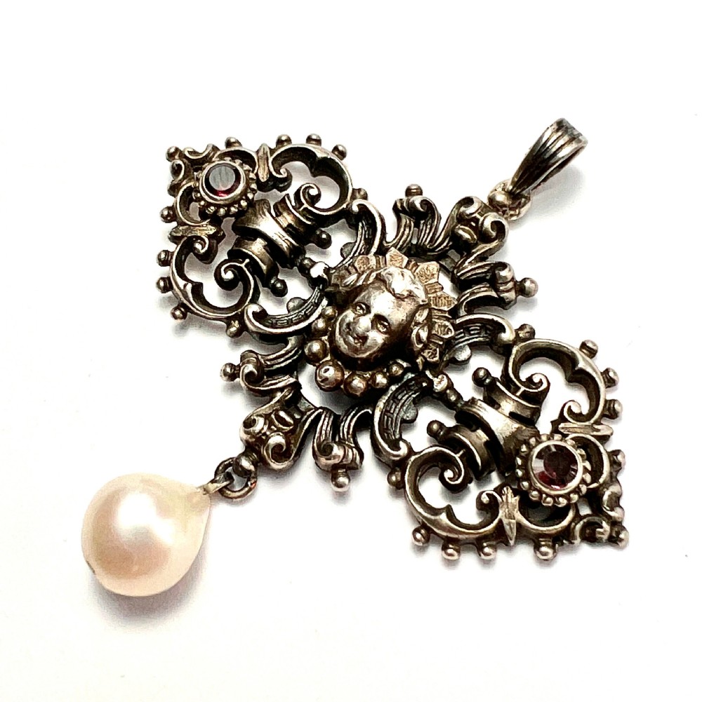 An antique silver pendant featuring scrollwork ornamentation, a central cherub mask, ruby-toned side accents, and a suspended baroque pearl drop. View IV