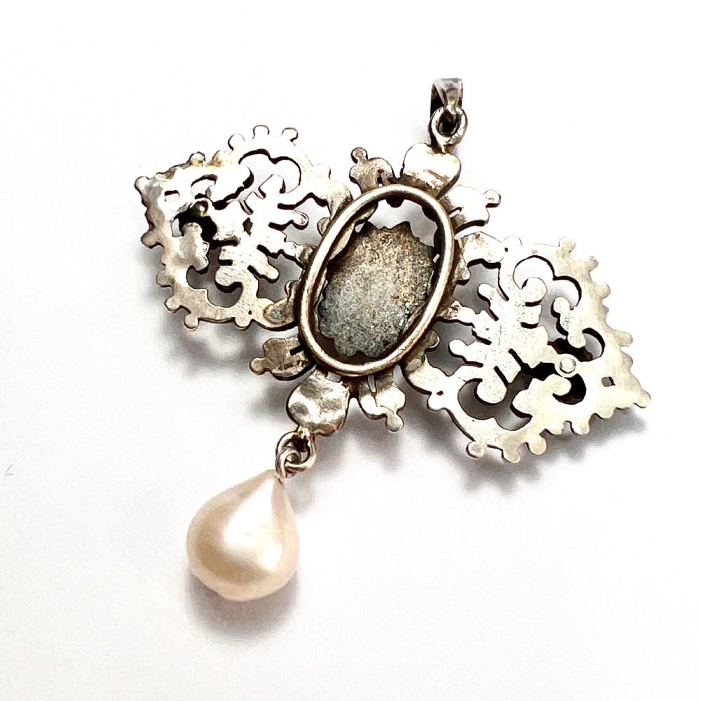 An antique silver pendant featuring scrollwork ornamentation, a central cherub mask, ruby-toned side accents, and a suspended baroque pearl drop. View V