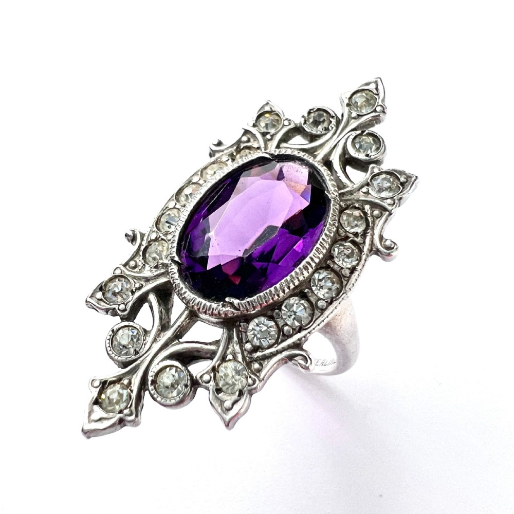A sterling silver cocktail ring featuring a central oval purple paste stone set within an openwork scroll setting studded with clear paste stones.