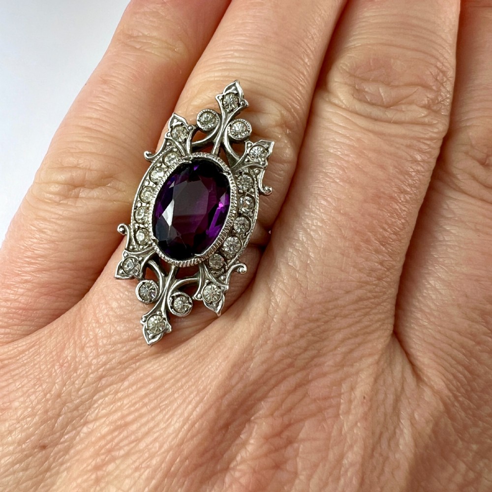 A sterling silver cocktail ring featuring a central oval purple paste stone set within an openwork scroll setting studded with clear paste stones. View VIII