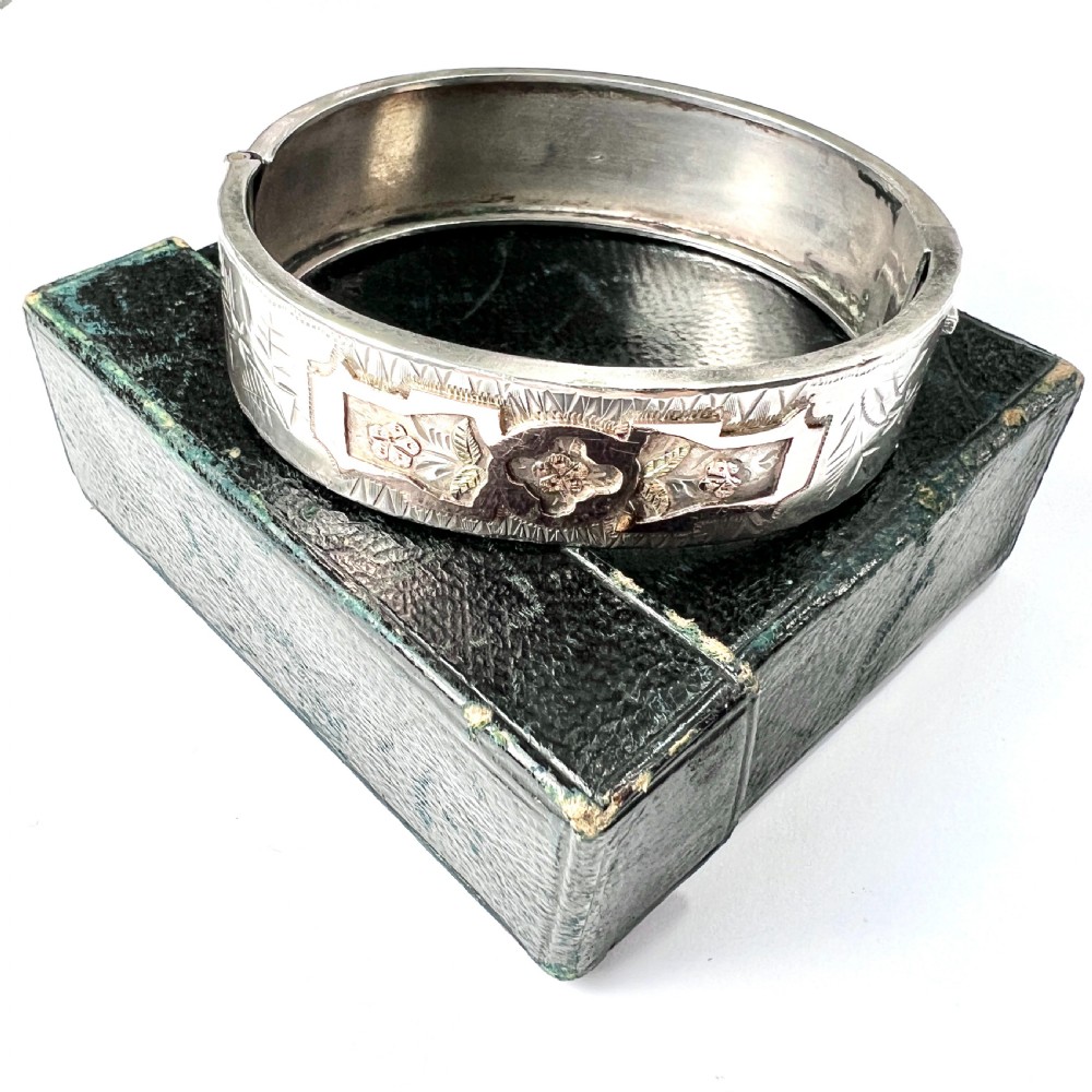 A Victorian sterling silver hinged cuff bangle decorated with engraved floral patterns and multi-tone gold overlays, resting on a green fitted presentation box.