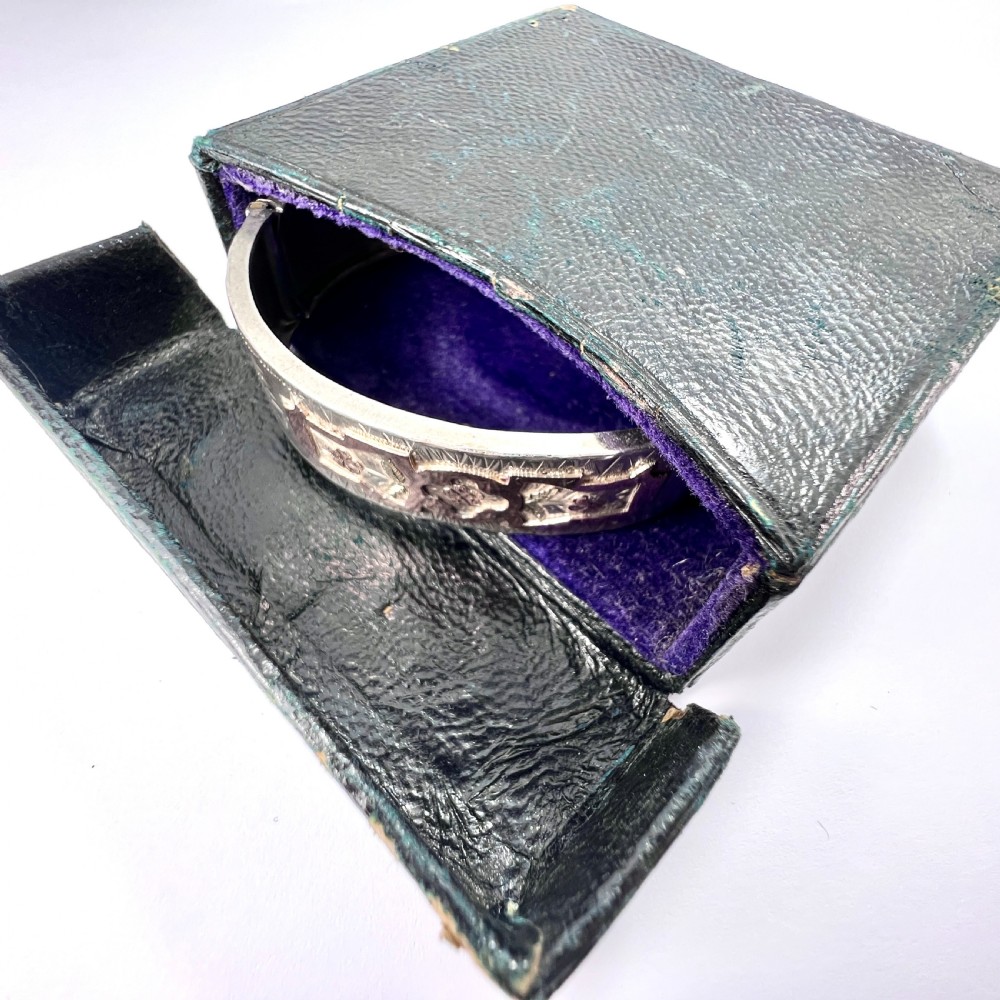 A Victorian sterling silver hinged cuff bangle decorated with engraved floral patterns and multi-tone gold overlays, resting on a green fitted presentation box. View II