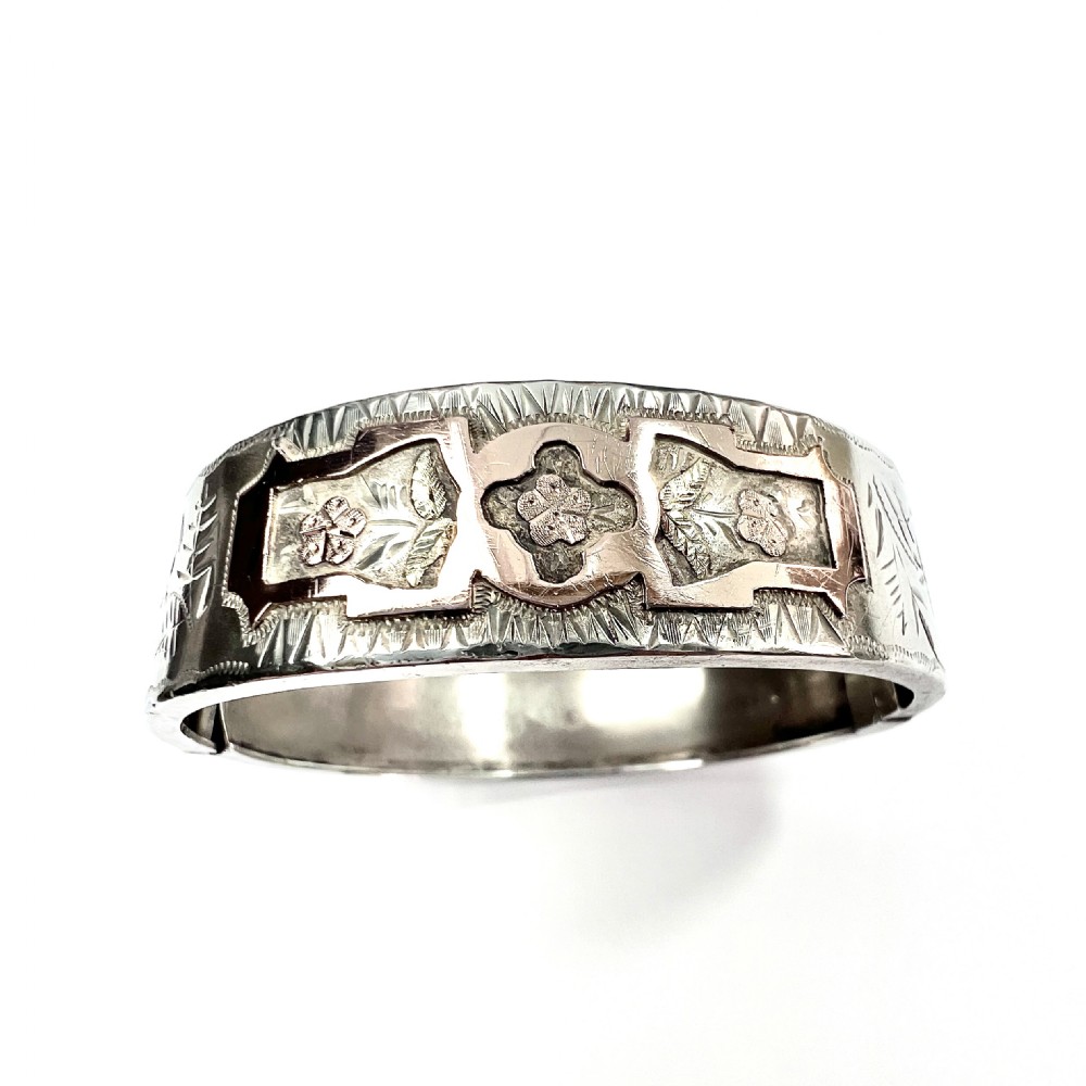 A Victorian sterling silver hinged cuff bangle decorated with engraved floral patterns and multi-tone gold overlays, resting on a green fitted presentation box. View III