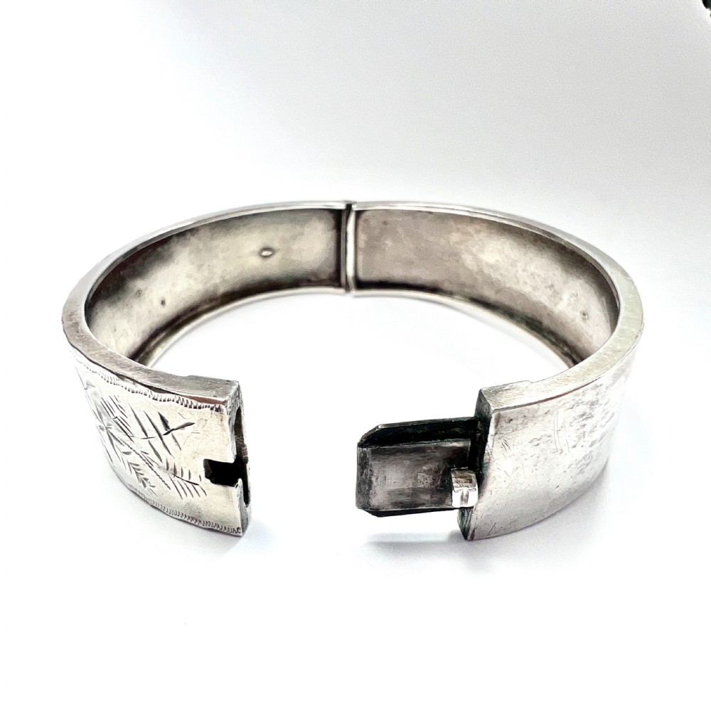 A Victorian sterling silver hinged cuff bangle decorated with engraved floral patterns and multi-tone gold overlays, resting on a green fitted presentation box. View V