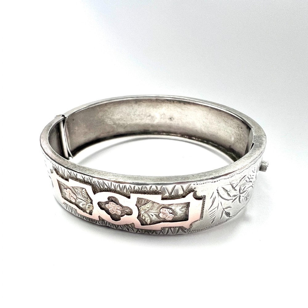 A Victorian sterling silver hinged cuff bangle decorated with engraved floral patterns and multi-tone gold overlays, resting on a green fitted presentation box. View VI