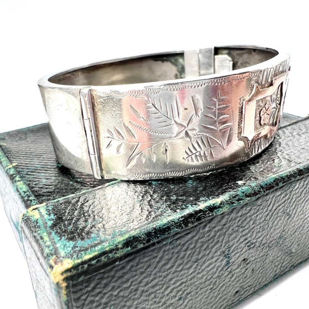A Victorian sterling silver hinged cuff bangle decorated with engraved floral patterns and multi-tone gold overlays, resting on a green fitted presentation box. View VII