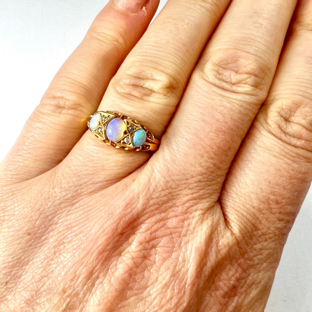 A Victorian 18ct gold ring featuring three cabochon opals separated by diamond accents within a carved scroll setting. View X