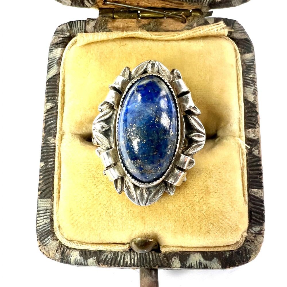 An Arts and Crafts style silver ring featuring a central oval blue lapis lazuli cabochon surrounded by chased silver leaf and ribbon motifs.