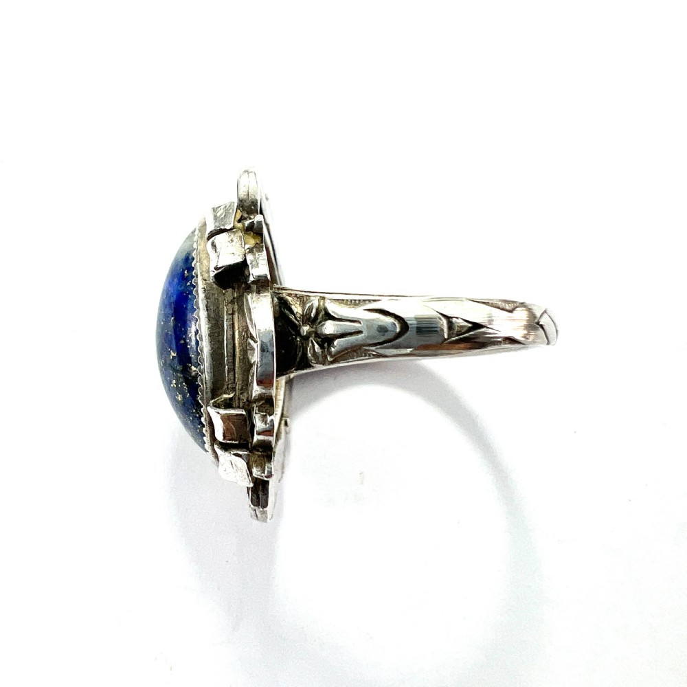 An Arts and Crafts style silver ring featuring a central oval blue lapis lazuli cabochon surrounded by chased silver leaf and ribbon motifs. View V