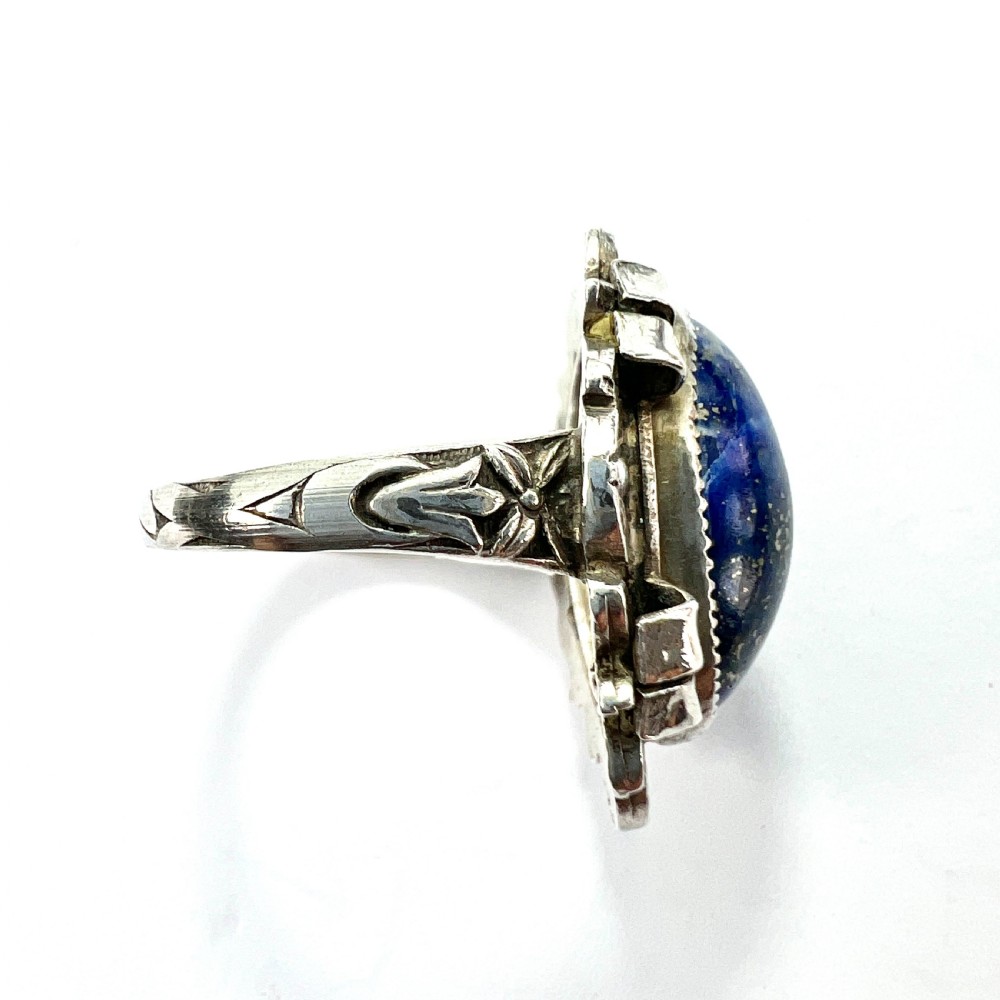 An Arts and Crafts style silver ring featuring a central oval blue lapis lazuli cabochon surrounded by chased silver leaf and ribbon motifs. View VI
