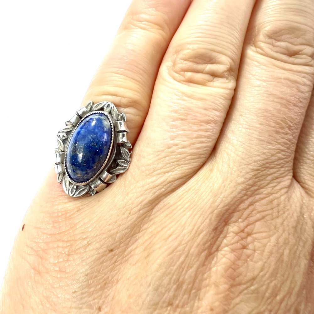 An Arts and Crafts style silver ring featuring a central oval blue lapis lazuli cabochon surrounded by chased silver leaf and ribbon motifs. View VIII