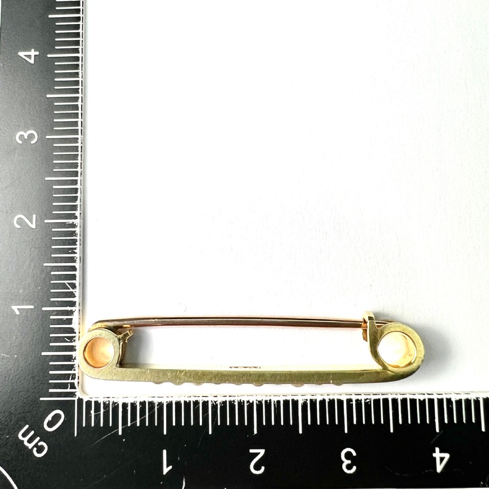 An antique 15ct yellow gold bar brooch designed as a safety pin, set with a row of seed pearls framed by black enamel detailing. View II