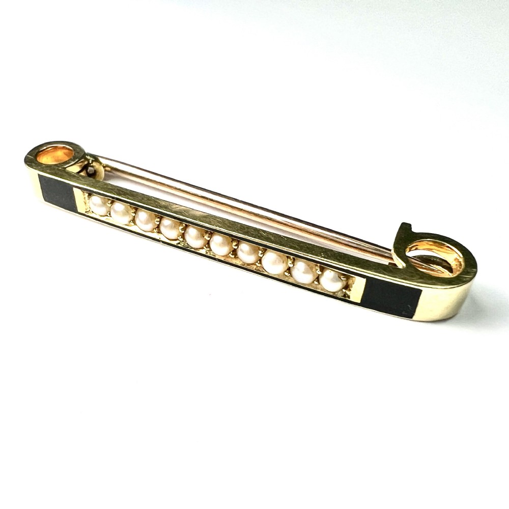 An antique 15ct yellow gold bar brooch designed as a safety pin, set with a row of seed pearls framed by black enamel detailing.