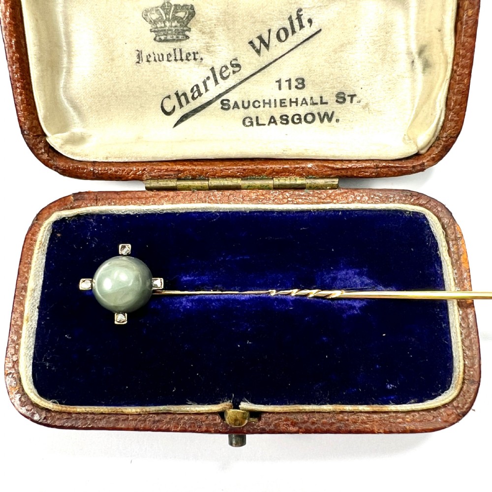 An antique 9ct gold stick pin featuring a central spherical green gemstone set with four small rose-cut diamonds, displayed inside an open velvet-lined leather jewellery box.
