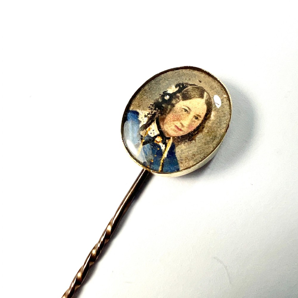 An antique gold stick pin featuring an oval miniature portrait painting of a lady in a blue dress with dark curls.