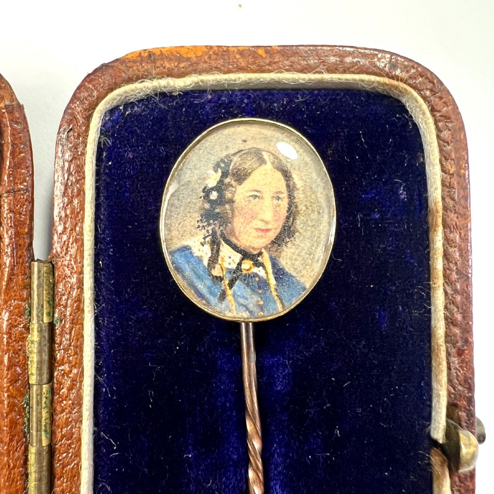 An antique gold stick pin featuring an oval miniature portrait painting of a lady in a blue dress with dark curls. View V