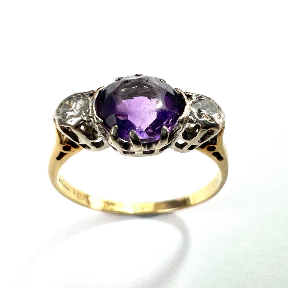 An antique 18ct yellow gold ring featuring a round purple central amethyst flanked by two diamonds in platinum settings. View IV