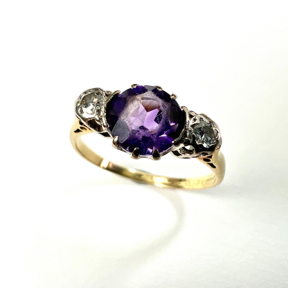 An antique 18ct yellow gold ring featuring a round purple central amethyst flanked by two diamonds in platinum settings. View V